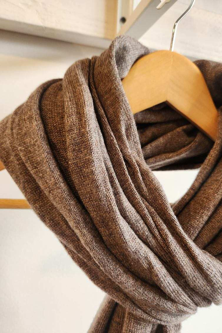 A cashmere scarf in a warm taupe-brown color, draped over a light wood hanger against a neutral off-white background.