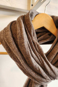 A cashmere scarf in a warm taupe-brown color, draped over a light wood hanger against a neutral off-white background.