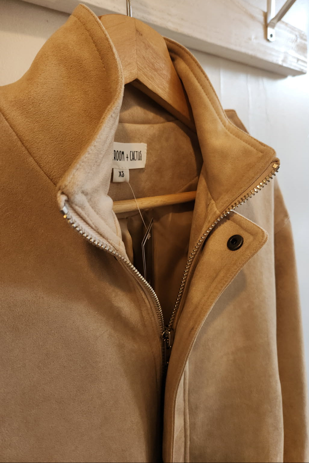 Brown suede jacket with a visible brand label on a hanger against a white wall.