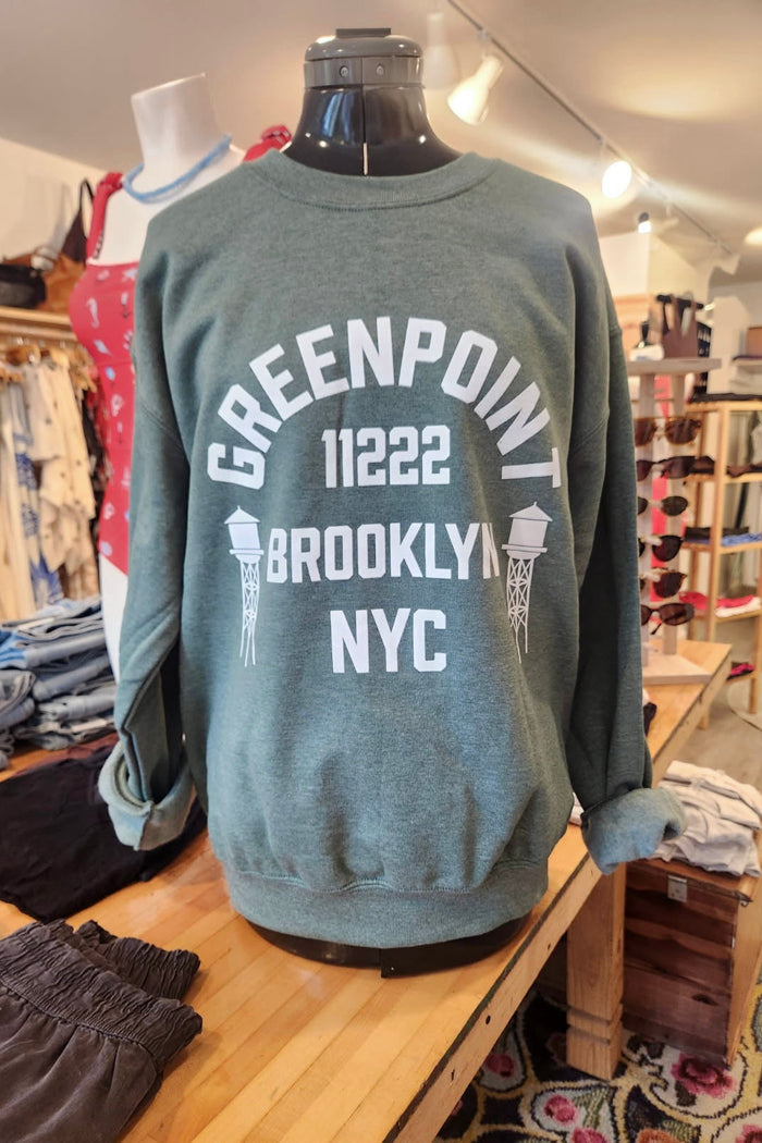 Green sweatshirt with 'Greenpoint 11222 Brooklyn NYC' text on a mannequin in a store setting.