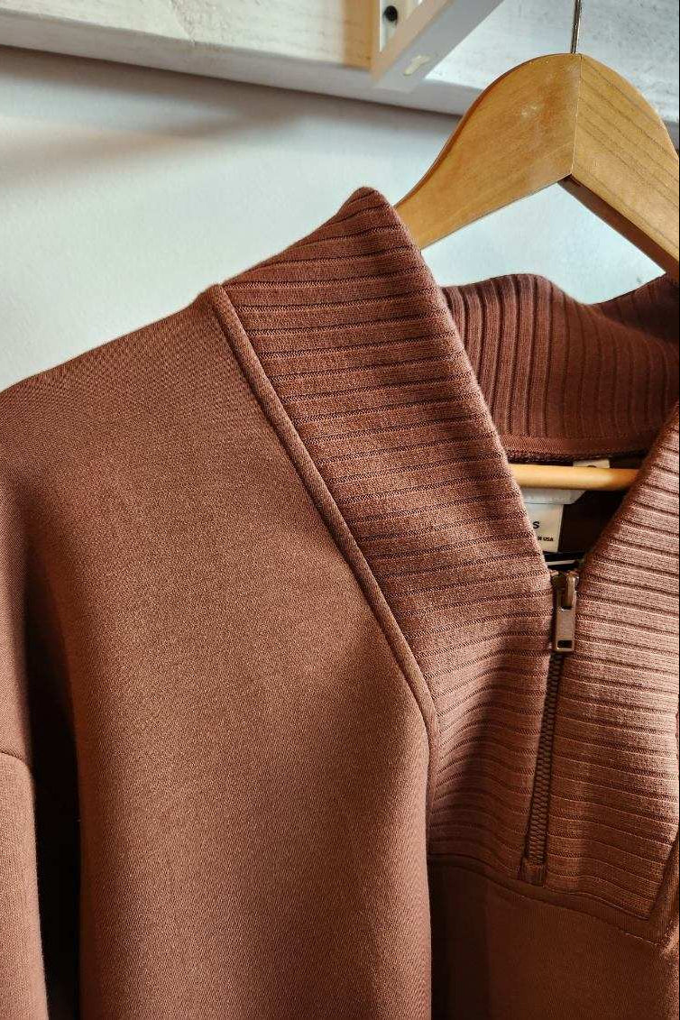 A close-up of a brown ribbed knit half-zip sweater hanging on a wooden hanger against a light wall.