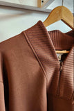 A close-up of a brown ribbed knit half-zip sweater hanging on a wooden hanger against a light wall.
