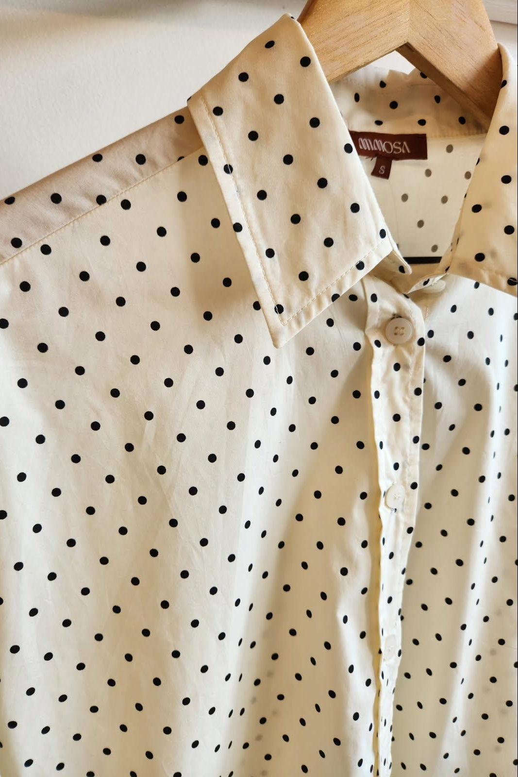 An off-white button-up shirt with a black polka dot pattern, hanging on a wooden hanger against a plain light background. 