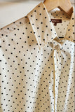 An off-white button-up shirt with a black polka dot pattern, hanging on a wooden hanger against a plain light background. 