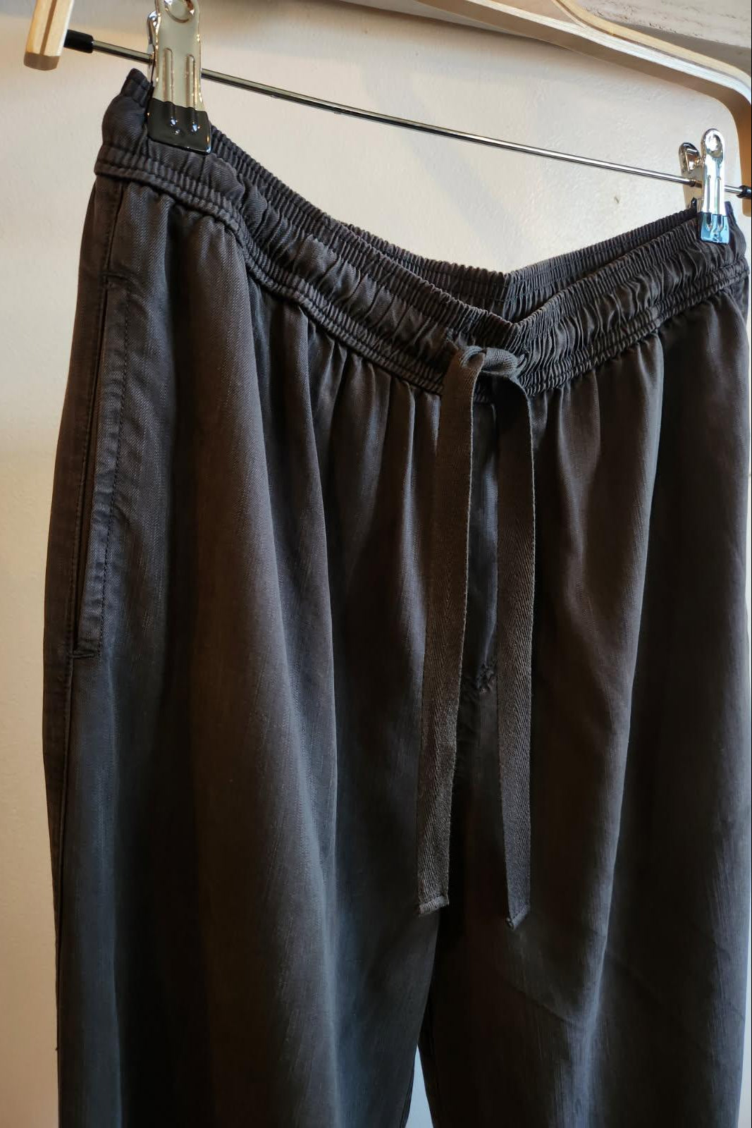 black drawstring pants hanging from a wooden hanger with metal clips against a plain wall.
