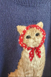 A close-up of a blue knit sweater featuring a beige cat motif on the chest, wearing a red patterned headband and red bow.