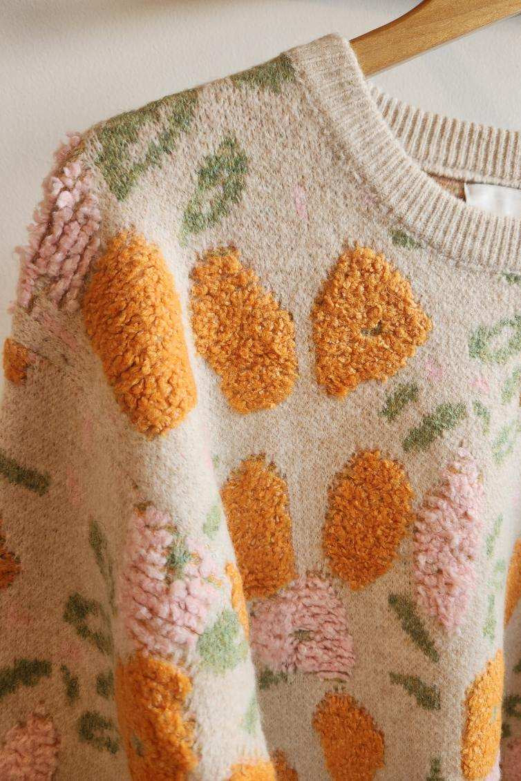 A close-up of a cream-colored knit sweater featuring raised, textured orange and pink floral motifs with green leaves, hanging on a wooden hanger.