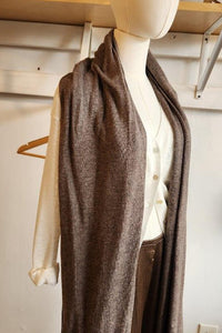 A long taupe-brown knit cardigan draped over a cream-colored long-sleeve top and brown trousers, displayed on a headless mannequin in a boutique setting.
