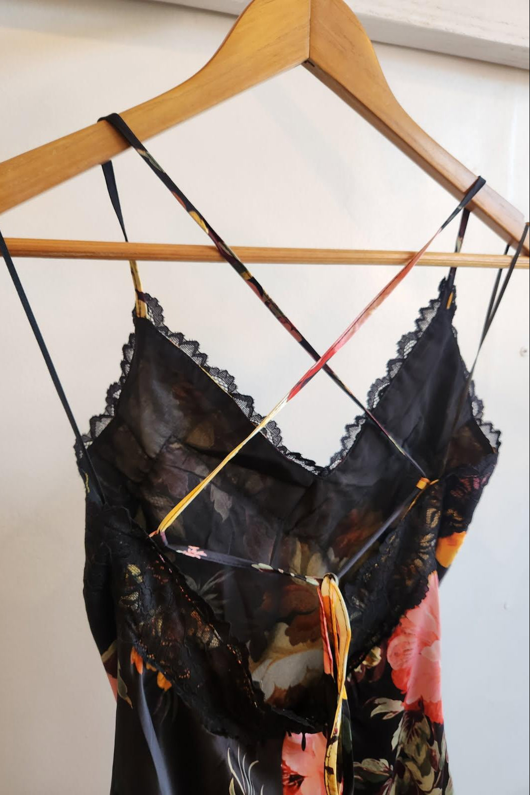 A sheer black floral slip dress with lace trim and thin multicolor patterned shoulder straps, hanging on a wooden hanger against a plain off-white wall.