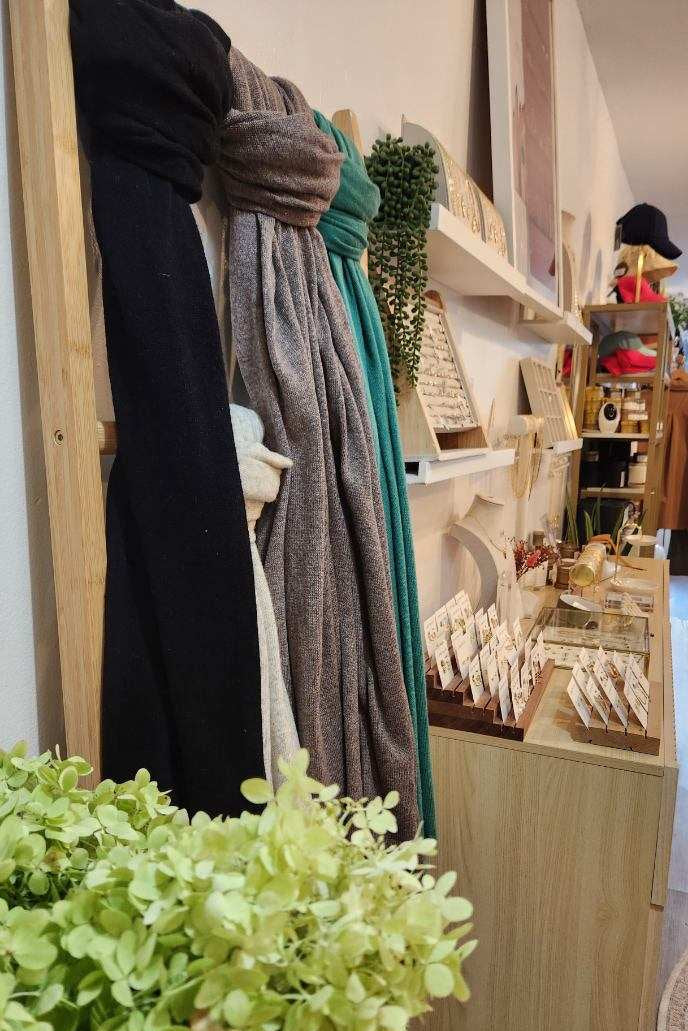 A boutique interior showing a wooden display rack with draped black and gray scarves, a teal wrap, and shelves holding small jewelry items and decor. A light wood counter with display stands is in the foreground, with a large green hydrangea in the lower left and a shop interior extending into the background.