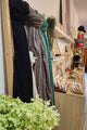 A boutique interior showing a wooden display rack with draped black and gray scarves, a teal wrap, and shelves holding small jewelry items and decor. A light wood counter with display stands is in the foreground, with a large green hydrangea in the lower left and a shop interior extending into the background.