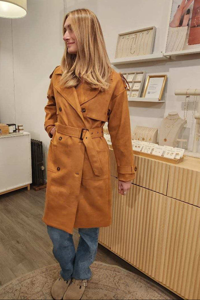 Woman wearing a tan coat standing in a store with wooden shelves and products.