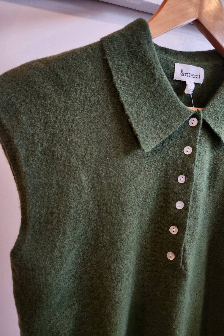 An olive green sleeveless knit polo shirt with a collar and button placket, hanging on a wooden hanger against a light wall. The brand label 'brmerei' is visible inside the collar.