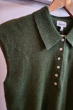 An olive green sleeveless knit polo shirt with a collar and button placket, hanging on a wooden hanger against a light wall. The brand label 'brmerei' is visible inside the collar.