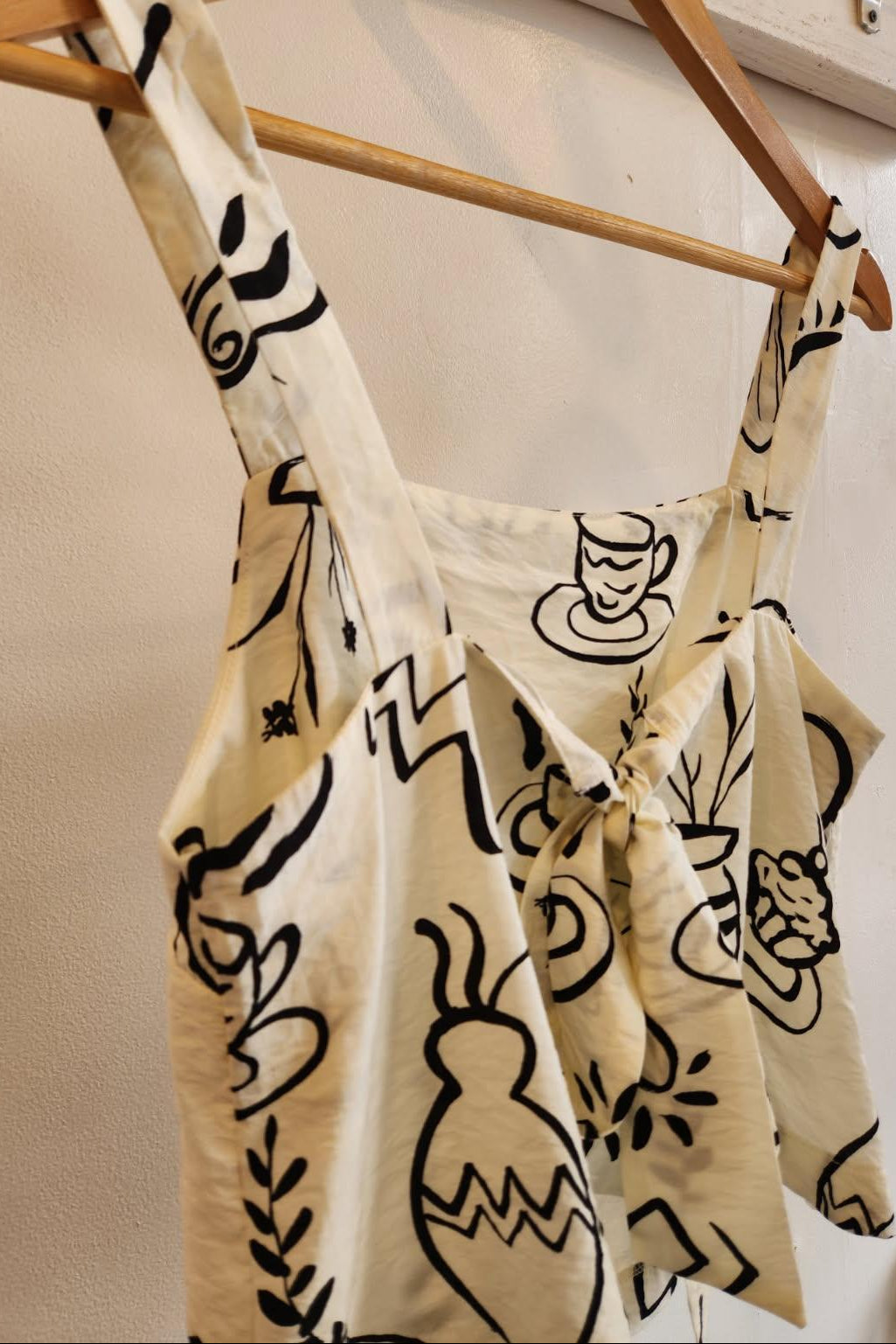 Beige tank top with black abstract patterns hanging on a wooden rack against a white wall.