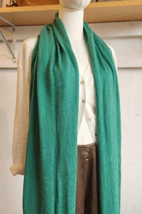 A long, solid teal-green knit scarf draped over a white mannequin torso, styled with a cream shirt and brown pants in a home-like interior setting.