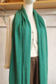 A long, solid teal-green knit scarf draped over a white mannequin torso, styled with a cream shirt and brown pants in a home-like interior setting.