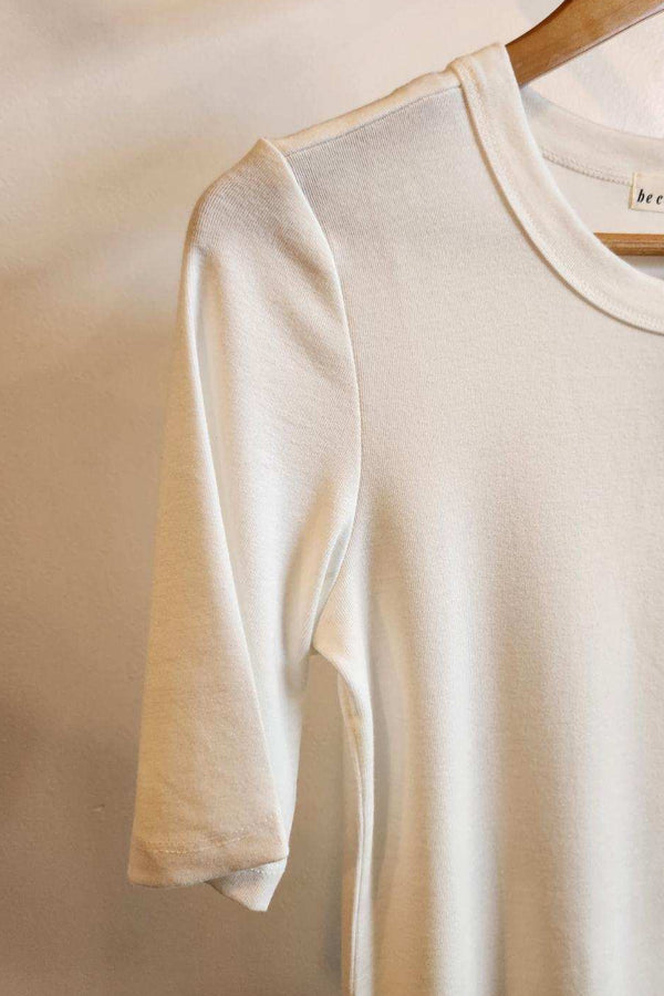 A cream-colored short-sleeve crewneck t-shirt hanging on a wooden hanger against a neutral beige wall.