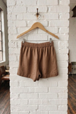Brown shorts hanging on a wooden hanger against a white brick wall.