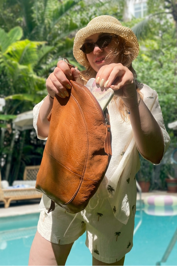 Person holding a brown leather bag by a pool with greenery in the background
