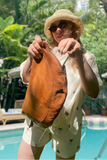 Person holding a brown leather bag by a pool with greenery in the background