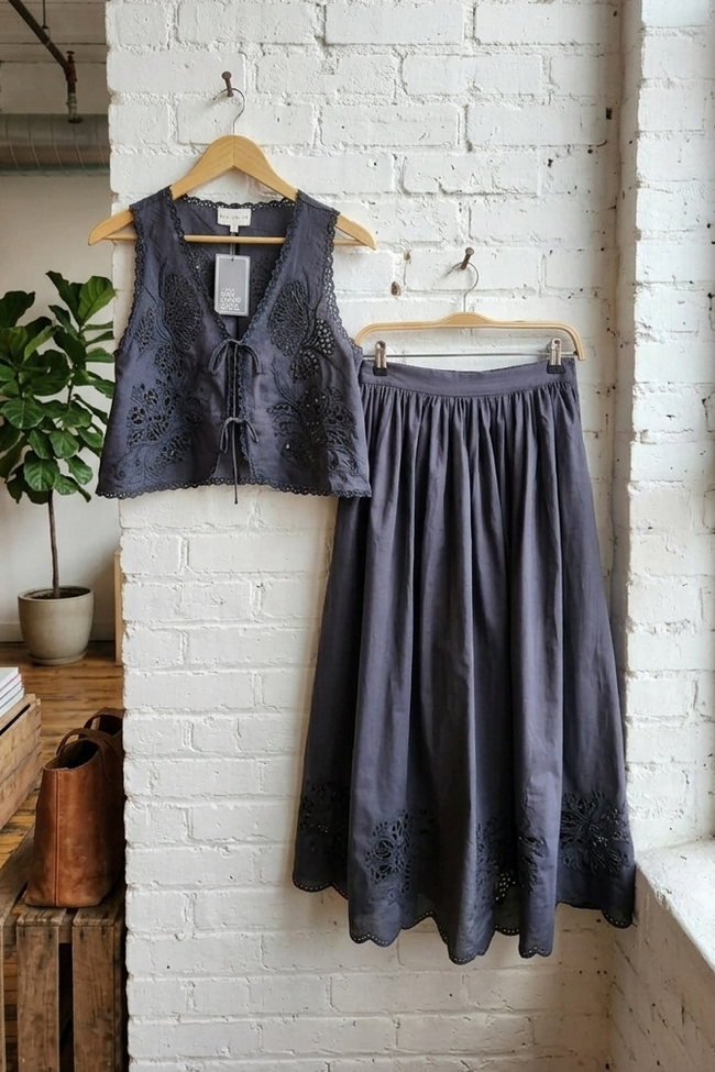 Vest and skirt hanging on a rack against a white brick wall in a room with a plant and wooden furniture.