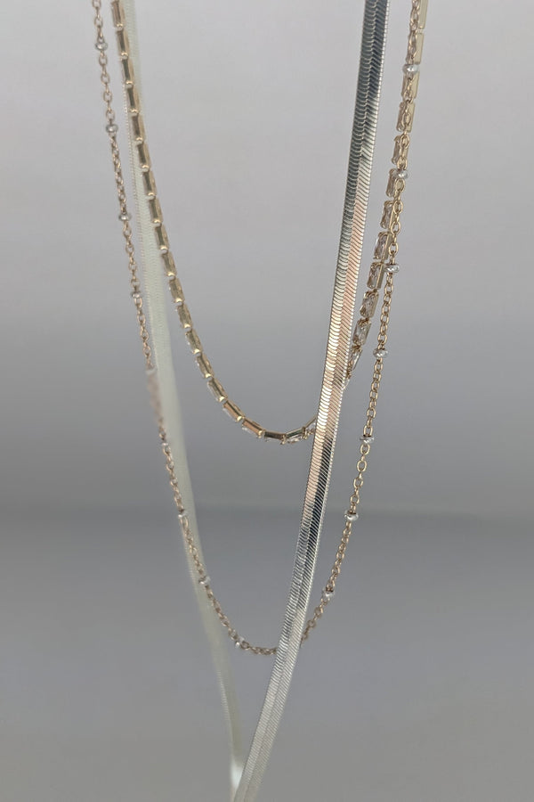 Three layered gold-tone and silver chain necklaces displayed on a neutral gray background.