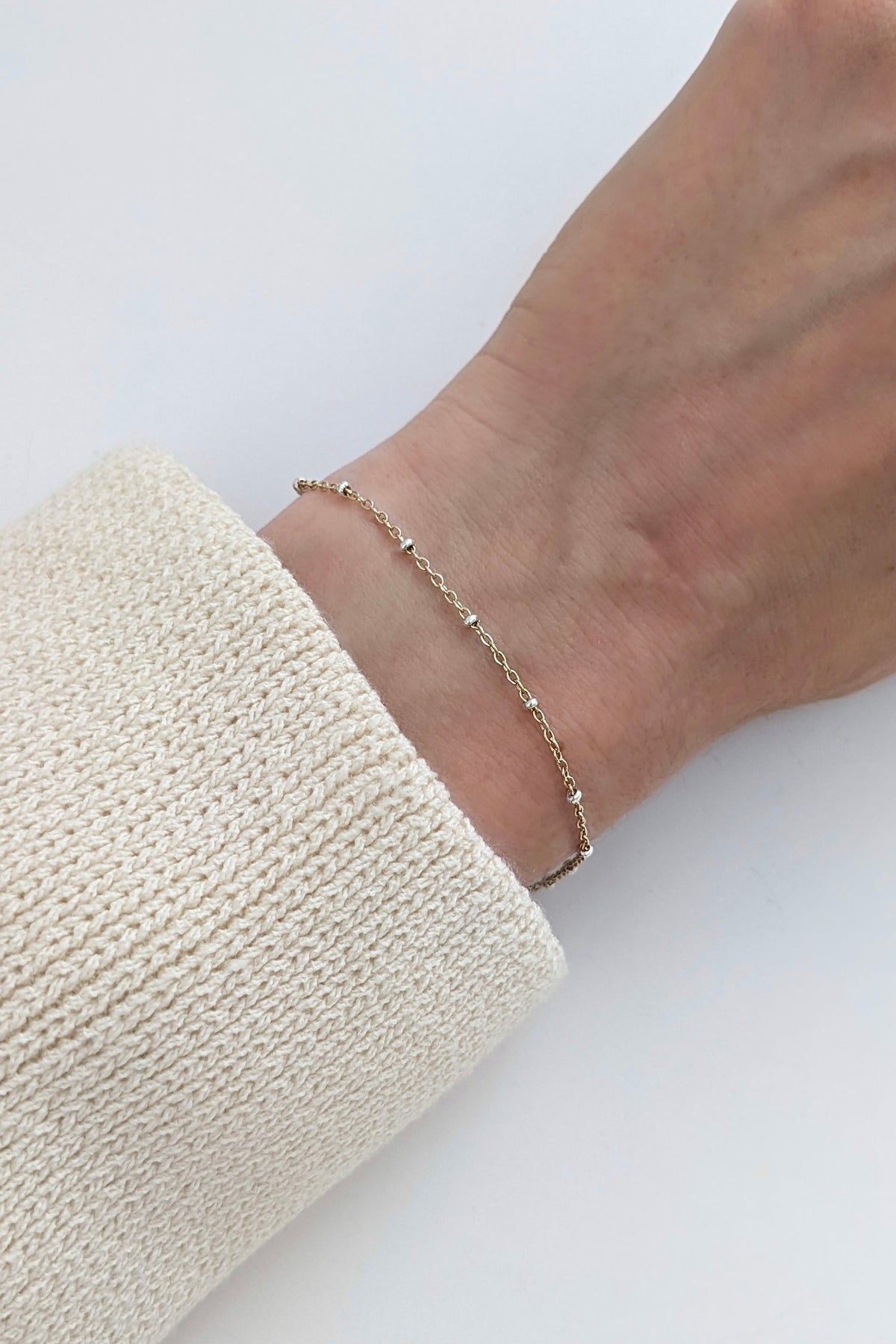 A delicate gold chain bracelet worn on a person's wrist, shown against a clean white background with a cream-colored knit sleeve visible.