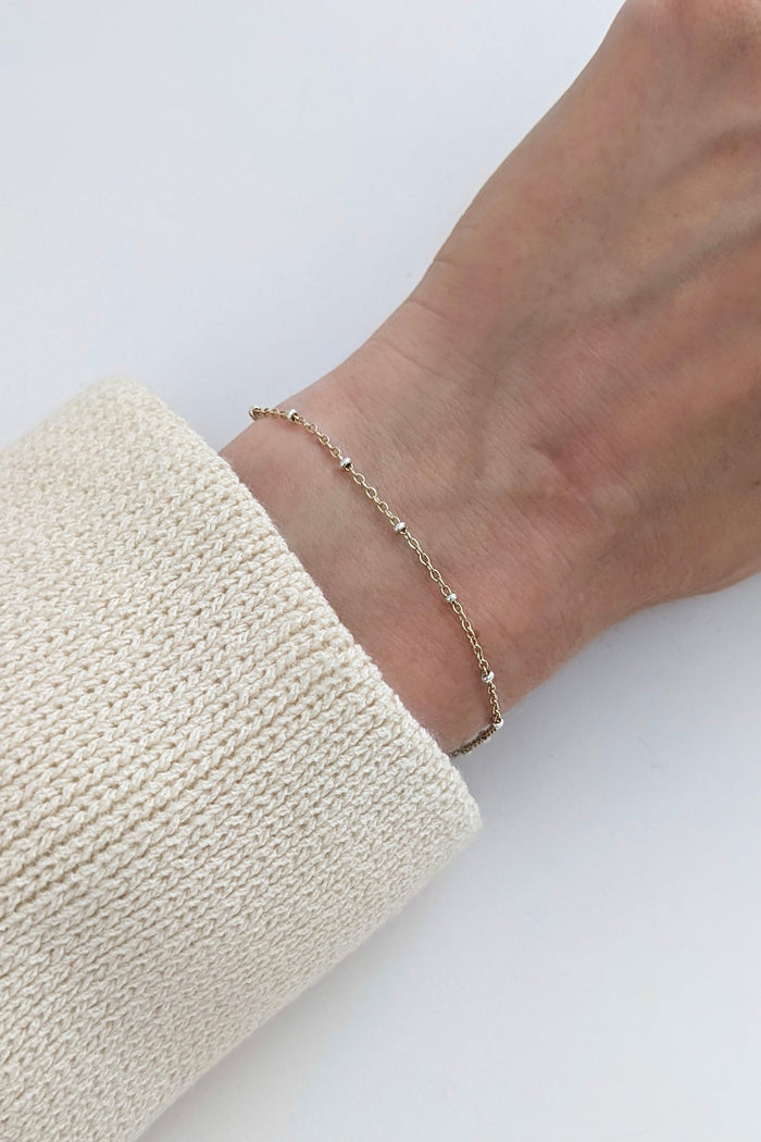 A delicate gold chain bracelet worn on a person's wrist, shown against a clean white background with a cream-colored knit sleeve visible.