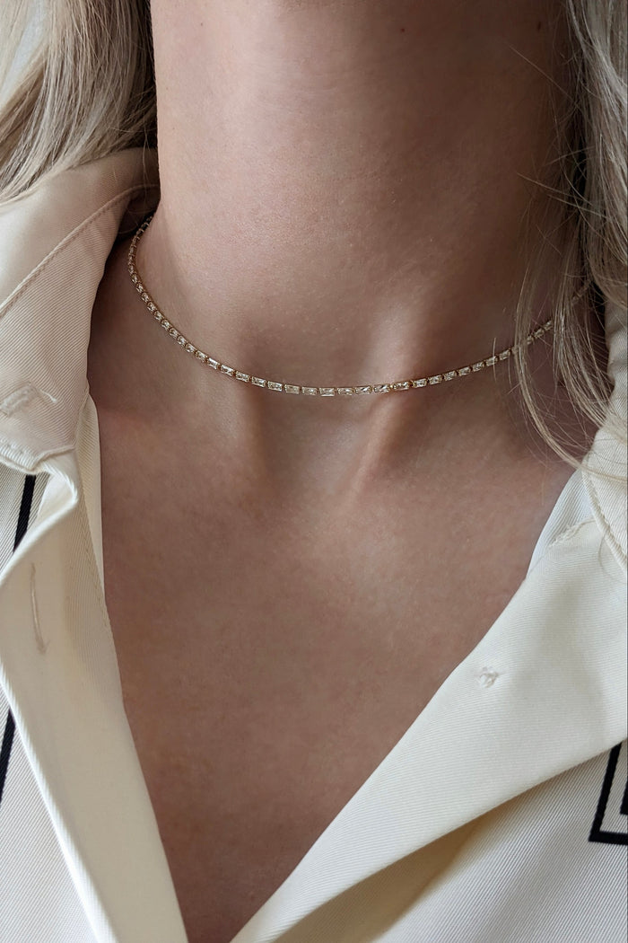 A close-up of a model's neck and collarbone wearing a delicate, thin gold chain choker necklace with small clear stones, paired with a cream-colored collared shirt.