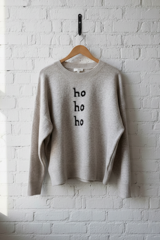 beige sweater with 'ho ho ho' text on a hanger against a white brick wall.