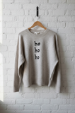 beige sweater with 'ho ho ho' text on a hanger against a white brick wall.