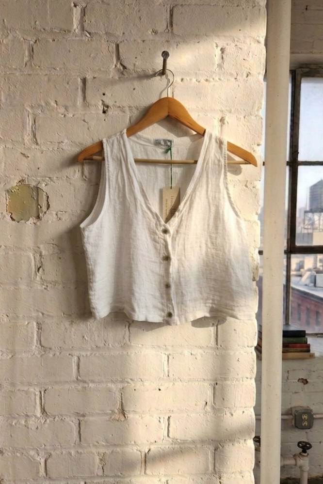 White sleeveless linen 
top on a wooden hanger against a white brick wall with a cityscape view.