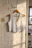 White sleeveless linen 
top on a wooden hanger against a white brick wall with a cityscape view.