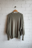 Green sweater hanging on a wooden hanger against a white brick wall.