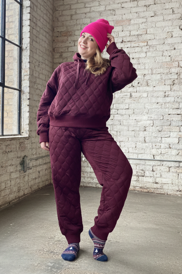 Person wearing a maroon quilted tracksuit and pink beanie in an urban setting with brick walls and large windows.