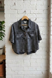A black washed short-sleeve button-up shirt with chest pockets hangs on a wooden hanger against a white brick wall in a loft-style room.