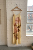 Yellow floral dress hanging on a wooden hanger against a white brick wall with a window in the background.