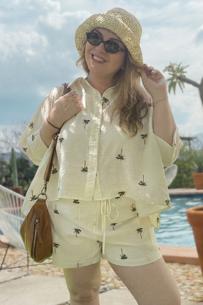 Woman in a light-colored outfit with a hat and sunglasses, standing by a poolside.