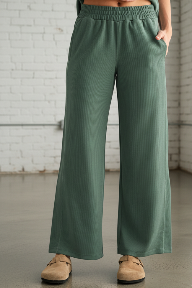 Person wearing green wide-leg pants in an indoor setting with a white brick wall.