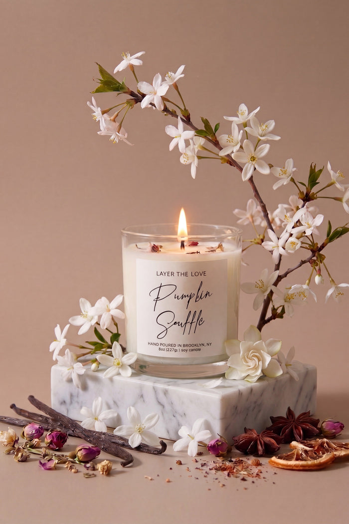 A lit glass jar candle labeled 'Purple Snuffle' by Layer The Love, styled on a white marble block with white blossoms, dried flowers, cinnamon sticks, and star anise against a warm beige background.