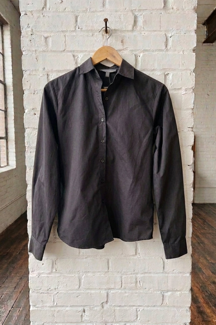 A black long-sleeve button-down shirt hanging on a wooden hanger against a white brick pillar in an industrial loft setting.
