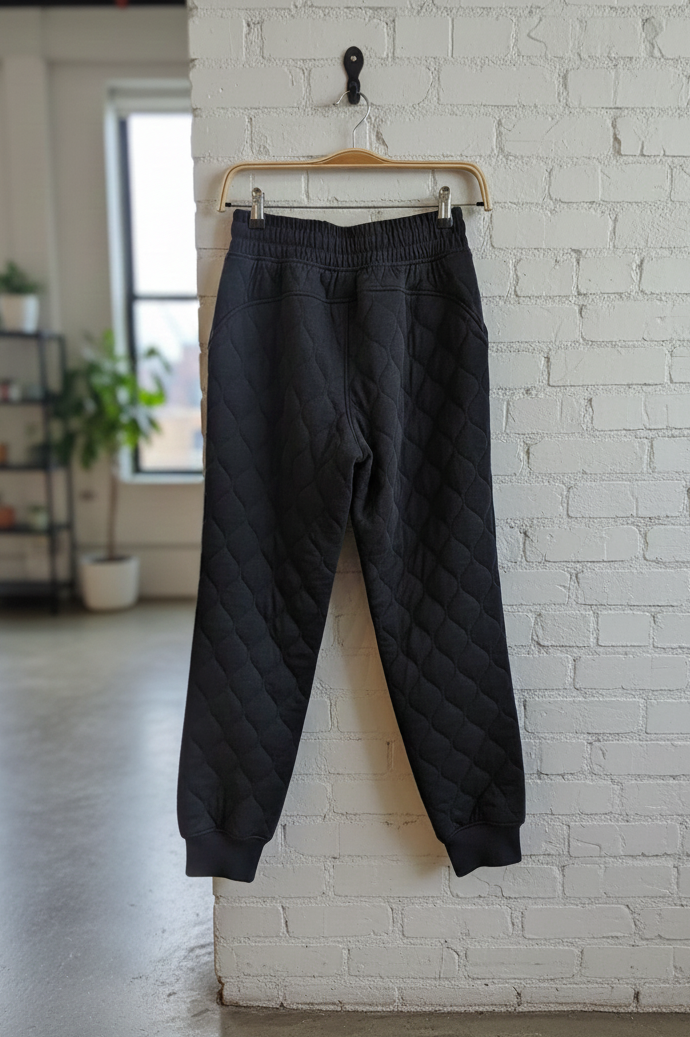 Black quilted pants hanging on a hanger against a white brick wall.