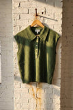 An olive green knit polo vest with a collar and three-button placket hangs on a wooden hanger from a hook on a white brick wall.