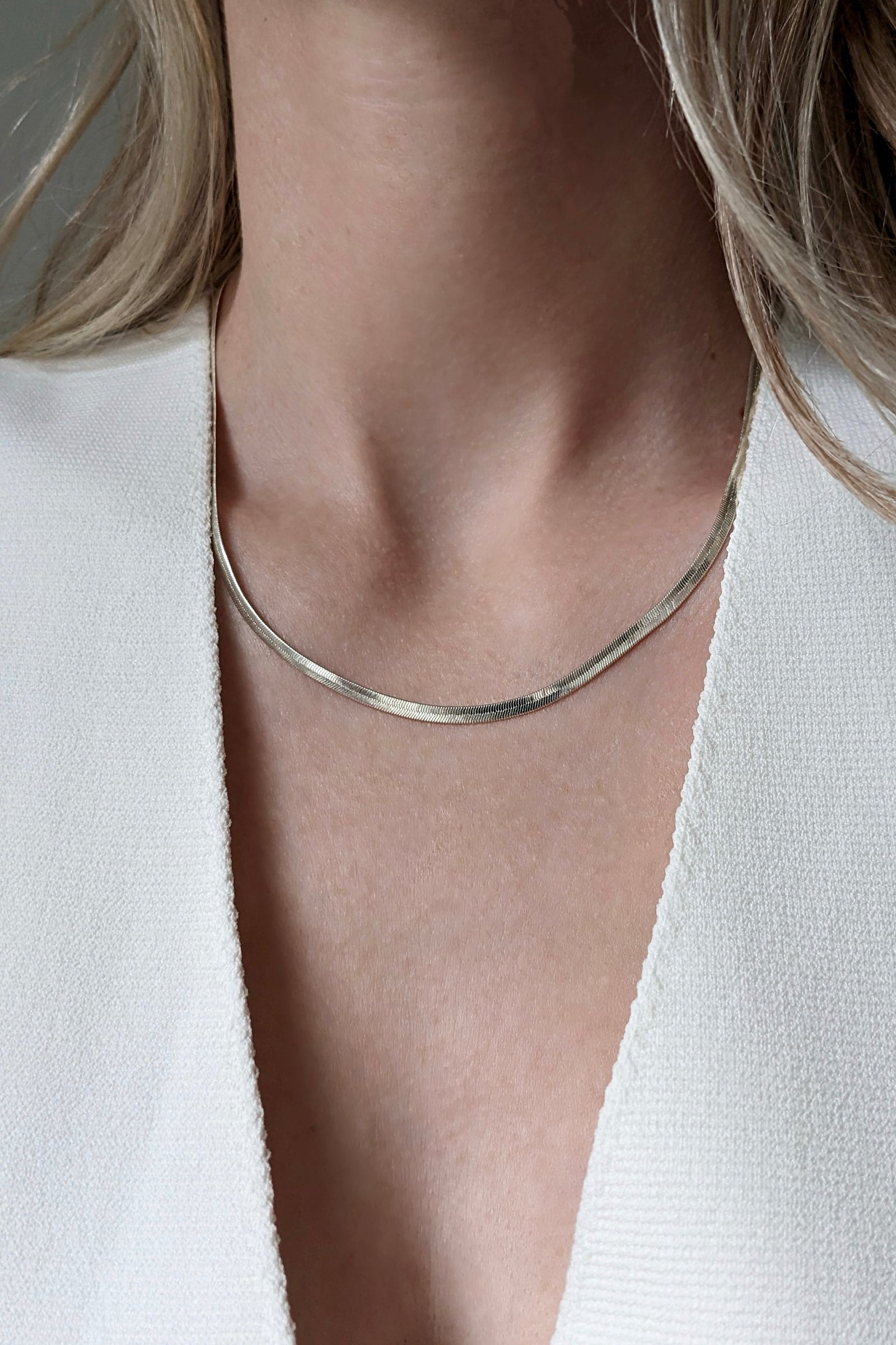 A close-up of a model's neck and chest wearing a thin, polished silver snake chain necklace and a white textured top.