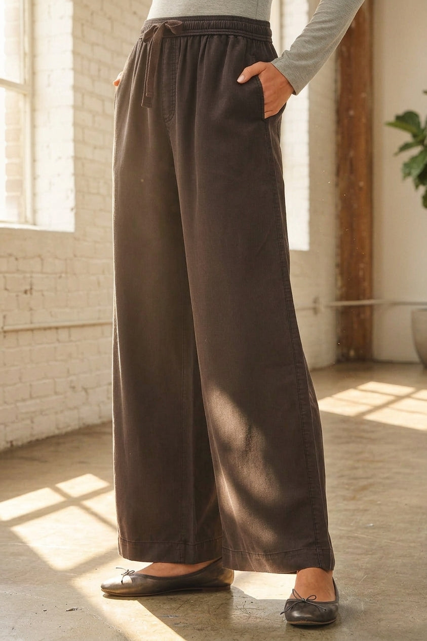 A person wearing black wide-leg drawstring pants with side pockets, paired with a light gray long-sleeve top and dark flats, standing in a sunlit room with a white brick wall and concrete floor.
