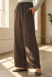 A person wearing black wide-leg drawstring pants with side pockets, paired with a light gray long-sleeve top and dark flats, standing in a sunlit room with a white brick wall and concrete floor.