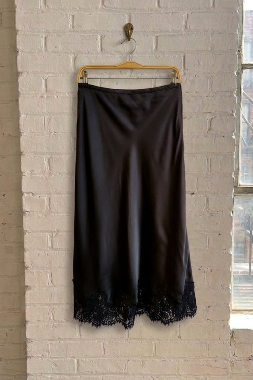 A black satin slip skirt with black lace trim at the hem, hanging from a wooden hanger on a white brick wall in a loft-style room with a window.