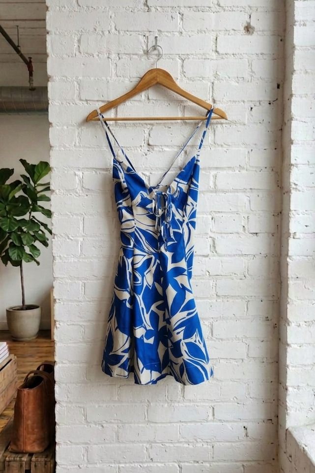 Blue and white floral dress hanging on a wooden hanger against a white brick wall.