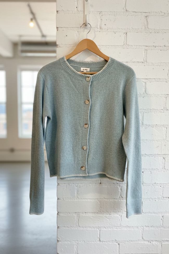 Light blue cardigan on a wooden hanger against a white brick wall.